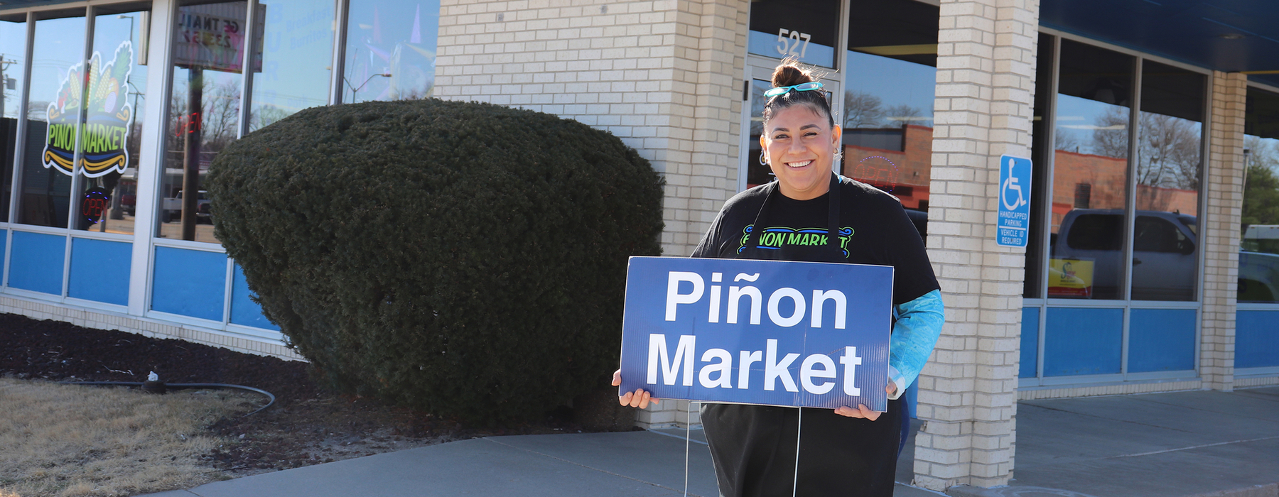 Latino woman stands outside grocery story holding a sign that says "Pinion Market"