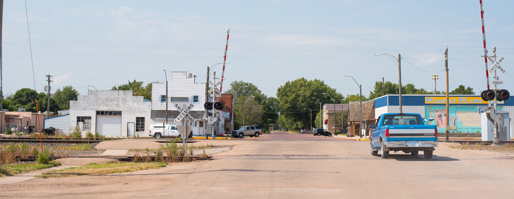 Blue and white truck crosses railroad tracks into a rural downtown with facades needing repair