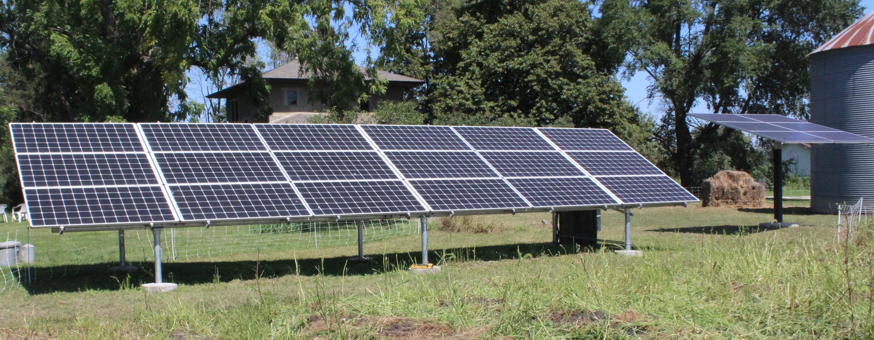 Solar panels sit on a farm with a grain bin and trees in the surround.