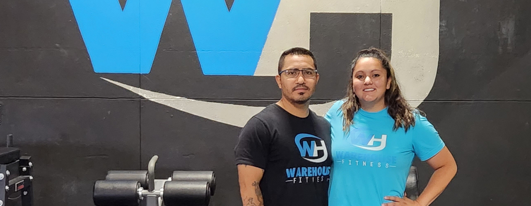 Man and woman wearing shirts that say "Warehouse Fitness" standing next to exercise equipment in front of a black wall with "WH"